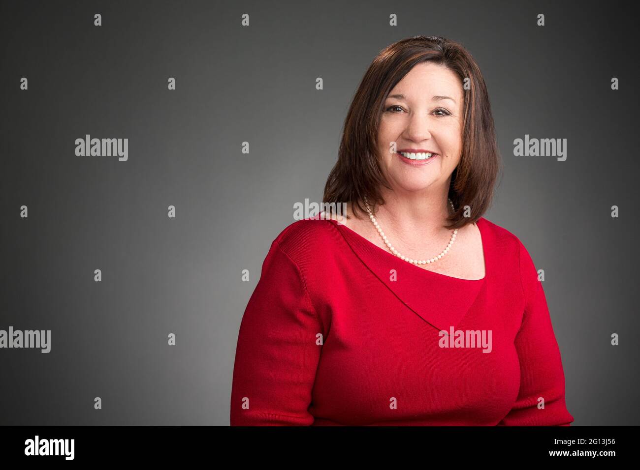 Eine lächelnde Frau in ihrem Firmenstudio – Porträt – Headshot. Grauer Hintergrund, horizontale Komposition. Eine glückliche Führungskraft in einem roten Kleid. Stockfoto