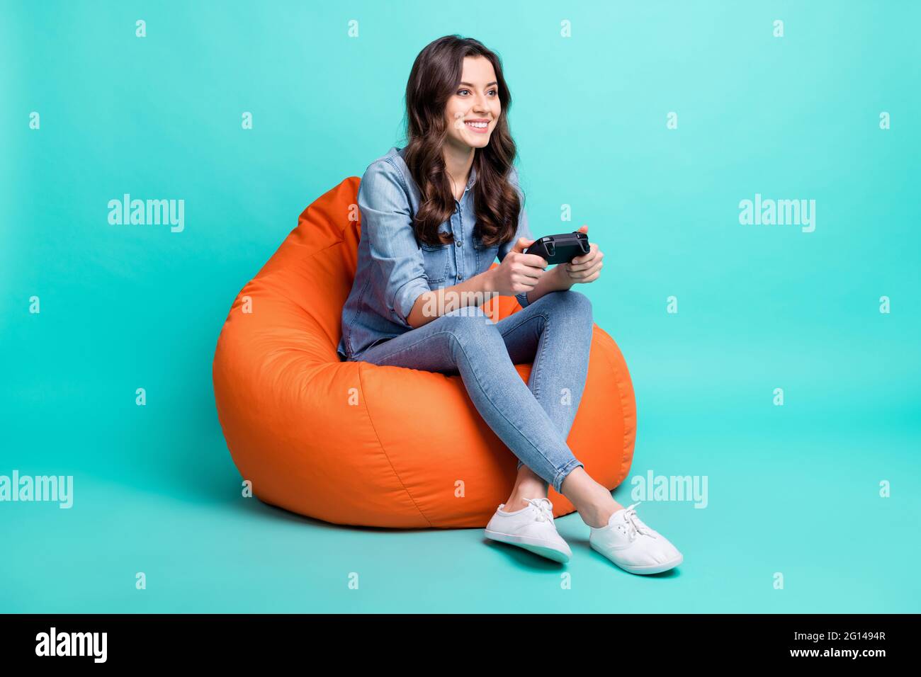 Ganzkörper-Foto von glücklichen schönen jungen Frau suchen leeren Raum sitzen Stuhl spielen Spiel isoliert auf blauem Hintergrund Stockfoto