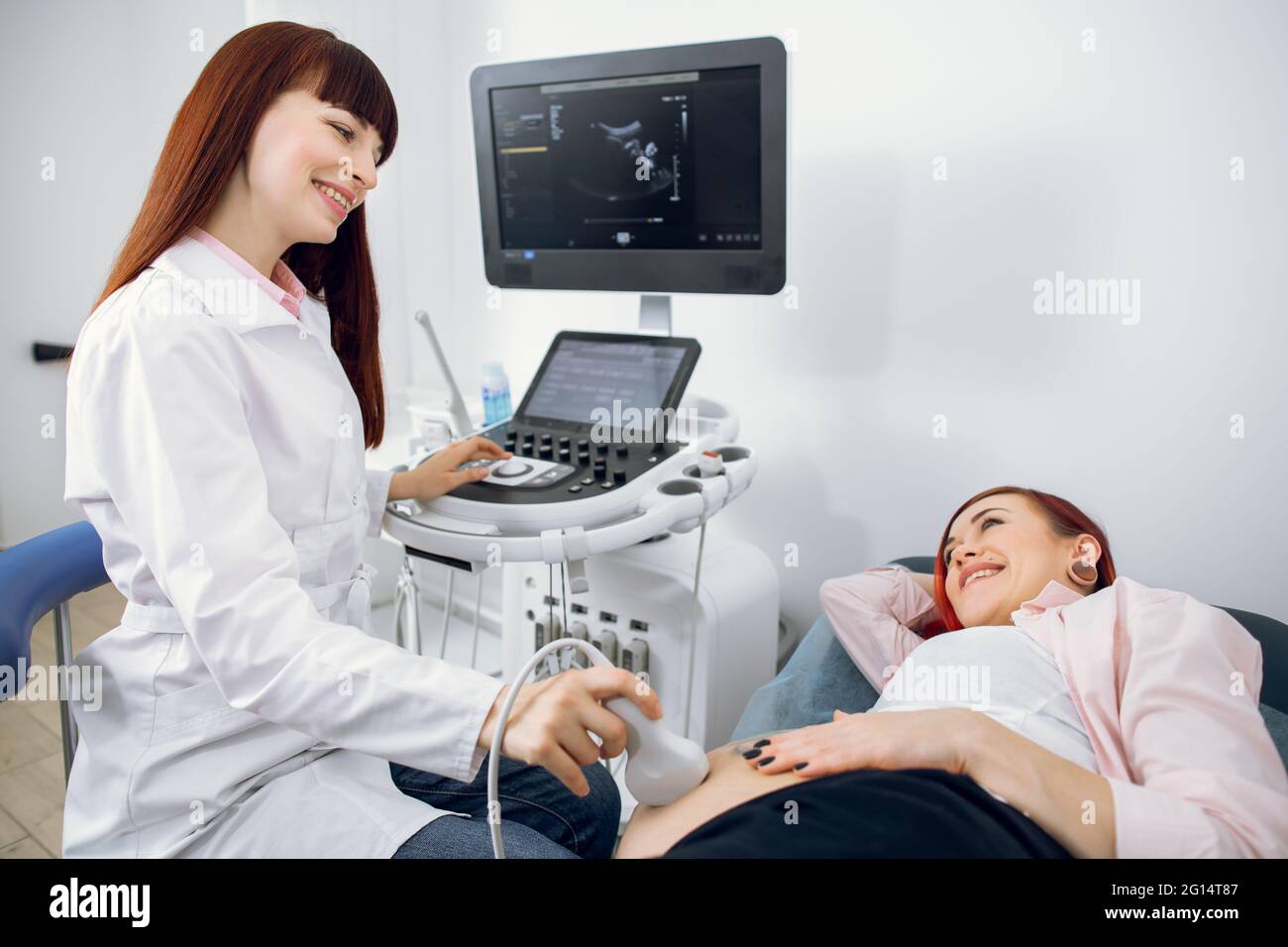 Eine ziemlich lächelnde Ärztin, die eine Ultraschalldiagnostik für ihre junge, glückliche, schwanger gebliebene Patientin durchführt. Glückliche, schwanger Dame genießt Ultraschall-Scan ihrer Zwillinge Babys Stockfoto