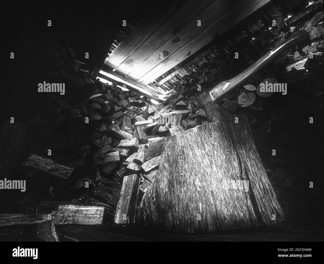 Holzschuppen mit bedrohlich wirkender Axt und großem Holzblock mit viel gestaltetem Feuerholz im Hintergrund. Lichteinfall im Holzschuppen Nahaufnahme Stockfoto