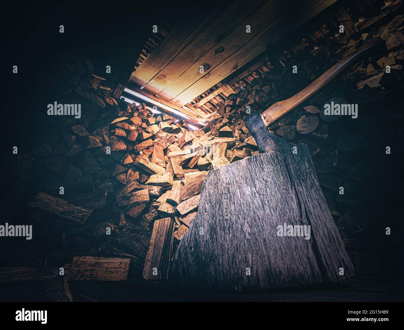 Holzschuppen mit bedrohlich wirkender Axt und großem Holzblock mit viel gestaltetem Feuerholz im Hintergrund. Lichteinfall im Holzschuppen Nahaufnahme Stockfoto