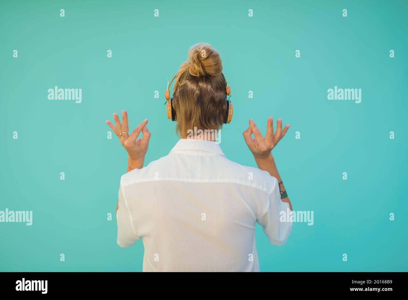 Rückansicht einer anonymen tätowierten Frau im schnurlosen Headset, die ein Lied hört, während sie auf aquamarinem Hintergrund Mudra der Weisheit zeigt Stockfoto