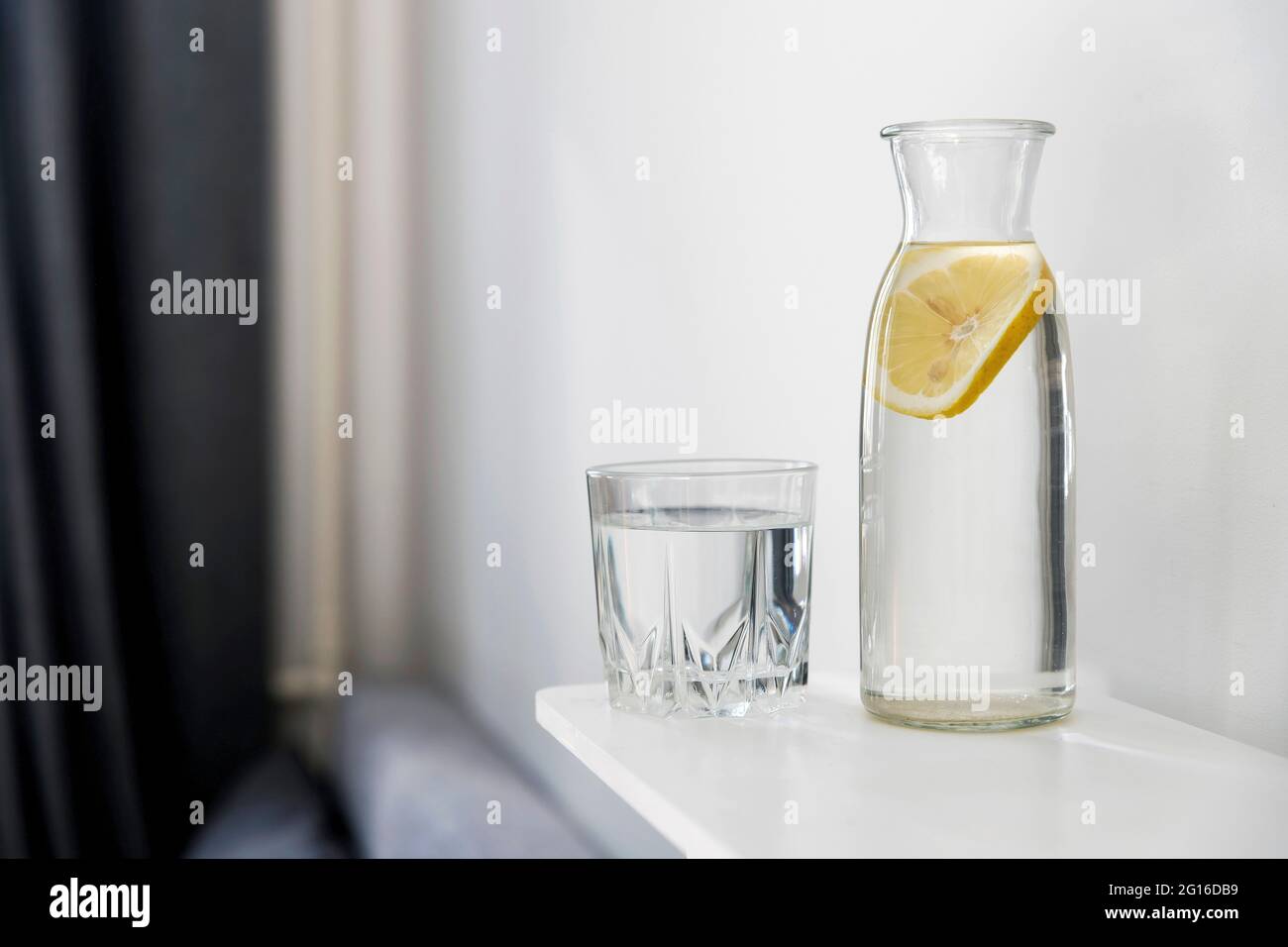 Ein erfrischendes Glas Wasser, eine Flasche mit einer Scheibe Zitrone im Inneren befindet sich in der Sommerhitze auf der Kommode Stockfoto