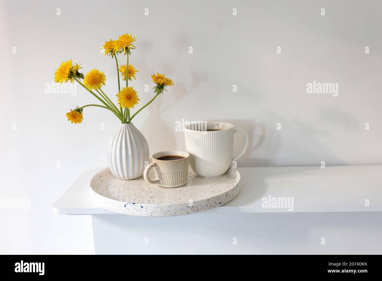 Ein Bouquet von Delelionen in einer weißen, geriffelten Vase und zwei Tassen in verschiedenen Größen mit Kaffee auf einem weißen Tisch. Frühstück. Speicherplatz kopieren Stockfoto