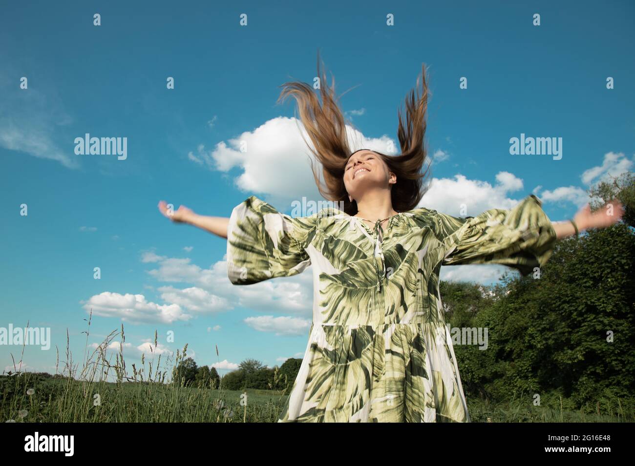 Junge Frau in Kleid im grünen Feld mit ausgestreckten Händen zu blauen Himmel und Sonne, fliegende Haare. Konzept von Glück, Freiheit und Unabhängigkeit Stockfoto
