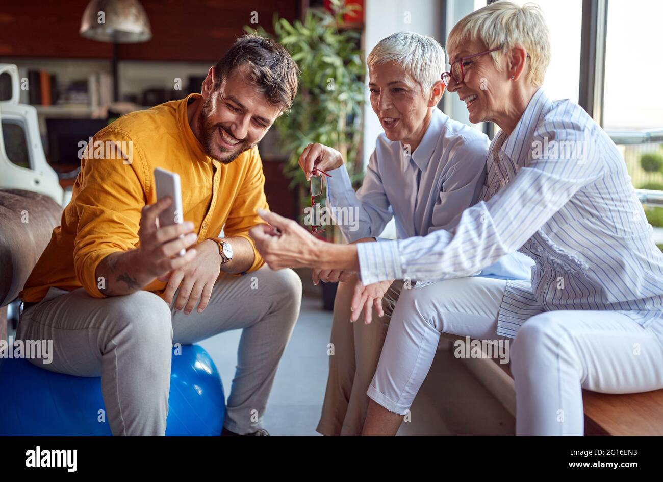 Ältere Geschäftsfrau kommentiert einen Smartphone-Content, den ihr junger männlicher Kollege ihnen zeigt, während sie in einer angenehmen Atmosphäre bei wor eine Pause einlegen Stockfoto