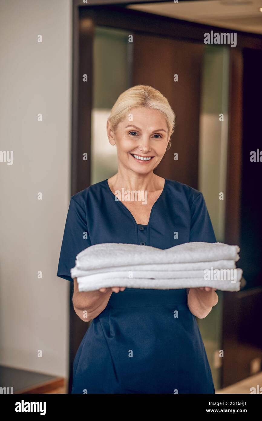 Lächelnde blonde Frau in Uniform hält saubere Handtücher Stockfoto