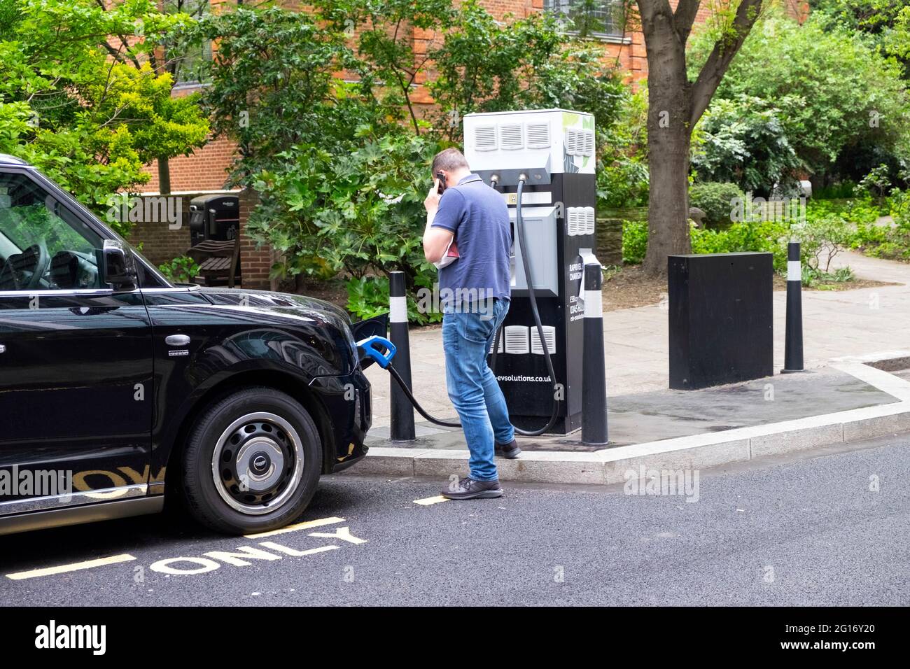Schwarzer Taxifahrer lädt sein E-Taxi an einer elektrischen Ladestation in der City of London, England, Großbritannien, KATHY DEWITT Stockfoto