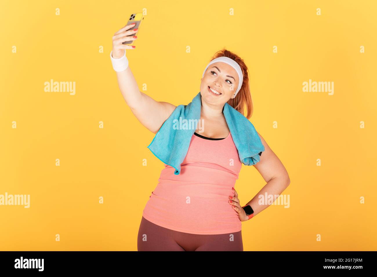 Fröhliche junge Plus-Größe Frau trägt sportliche Outfit und Handtuch nach dem Training hält Smartphone nimmt Selfie, während auf gelbem Hintergrund stehen. Stockfoto