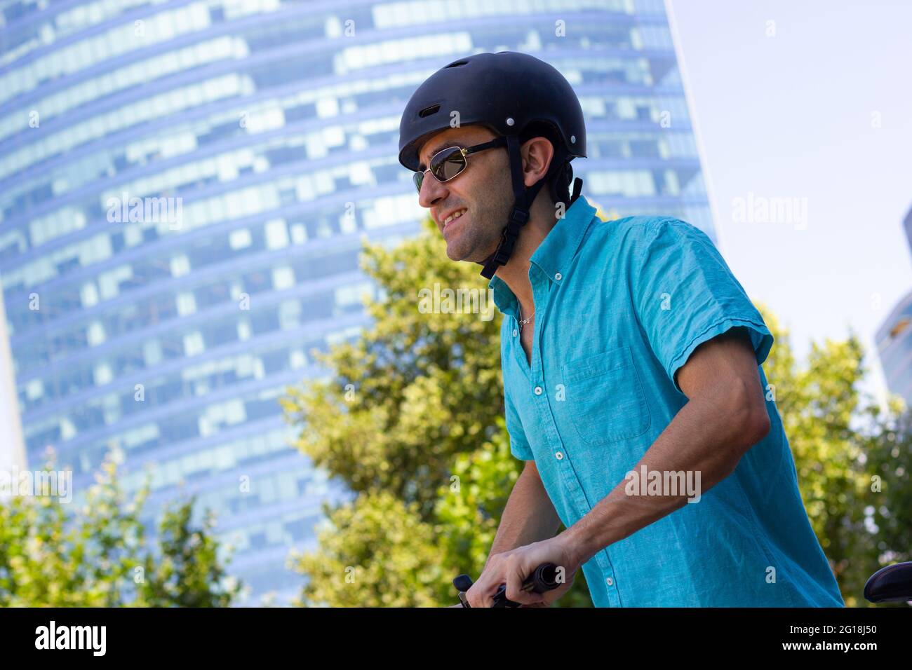 Lächelnder Mann mit Helm, der am sonnigen Tag in der Stadt Fahrrad fährt. Alternative Pendelanfahrtsmöglichkeit im Uban-Bereich mit Skyline im Hintergrund Stockfoto