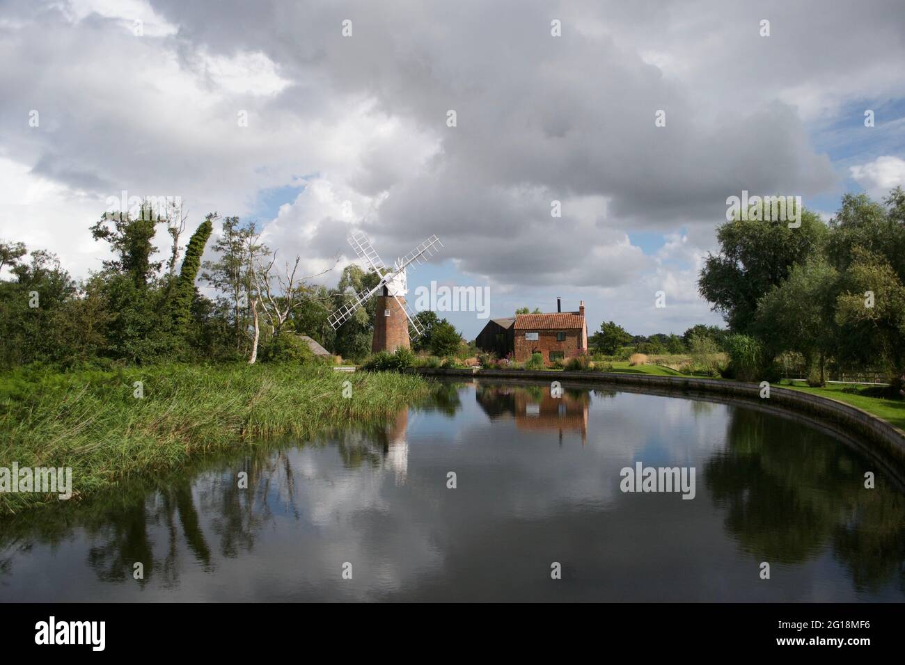 Eine traditionelle Windmühle (Windpumpe) und hübsches Häuschen an einer Flussbiegung in den Norfolk Broads, England, Großbritannien. Perfekte Reflexion unter dramatischem sto Stockfoto