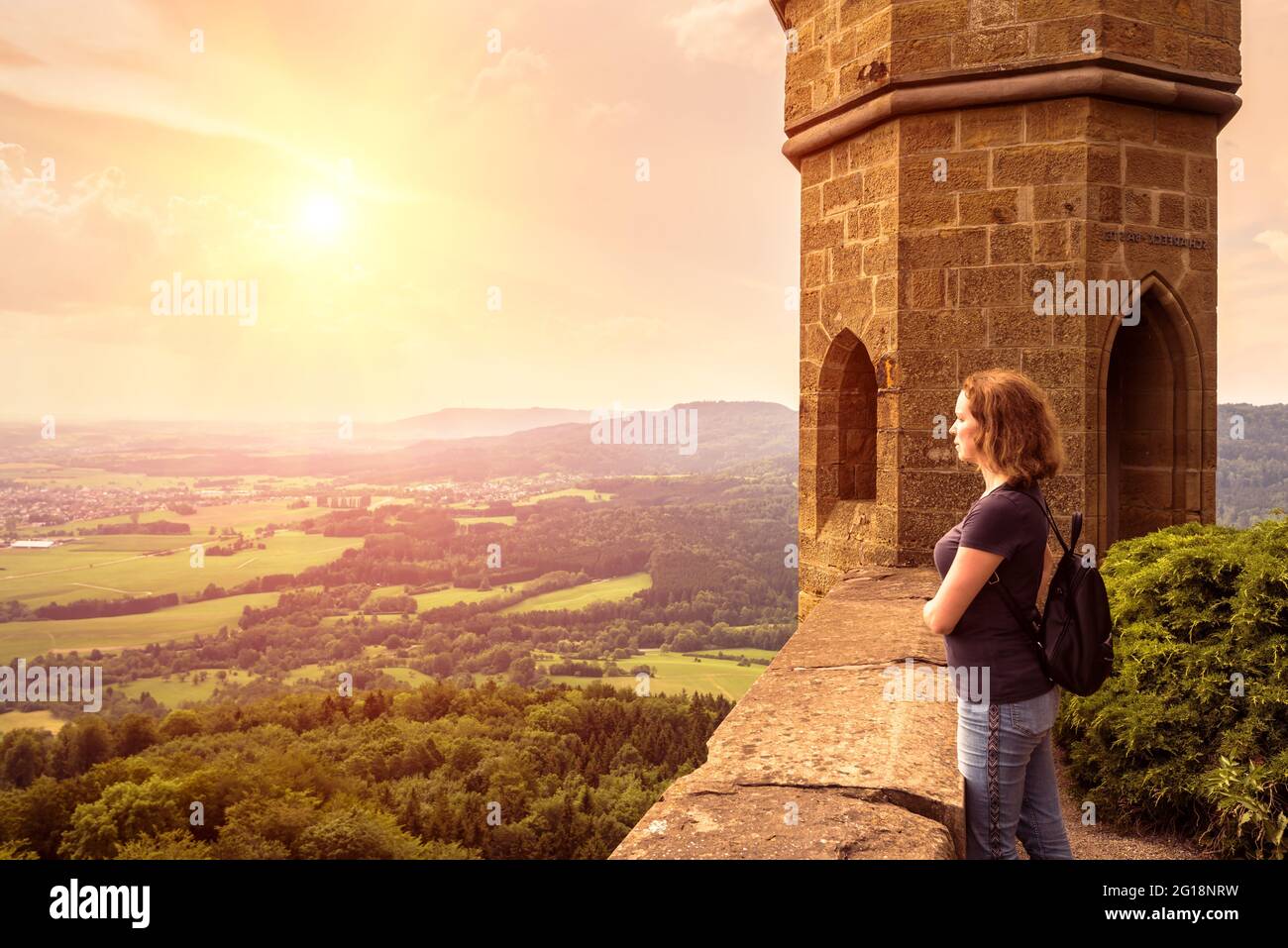 Junge Frau bewundert Landschaft bei Sonnenuntergang auf Schloss Hohenzollern, Deutschland. Dieser Ort ist eine Touristenattraktion in der Nähe von Stuttgart. Landschaftlich schöner Blick auf das sonnige Tal Stockfoto