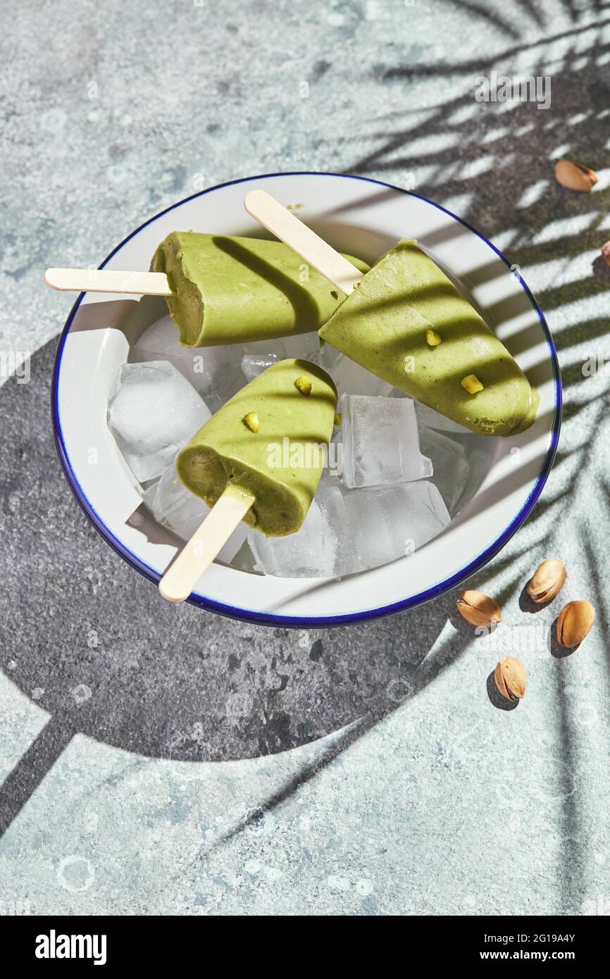 Gefrorener hausgemachter Pistazien-Popsicle in einer Eisschüssel auf grauem Hintergrund mit Palmenblatt-Schatten bei hartem Licht. Erfrischender Fruchteis, gefrorener grüner Saft auf Stockfoto