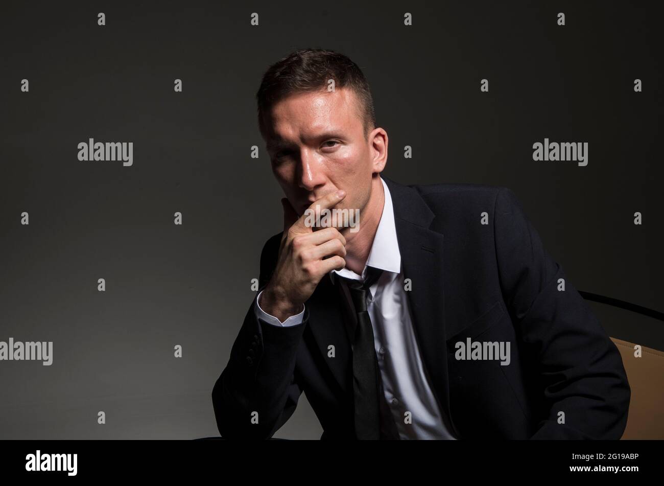 Porträt eines nachdenklichen, selbstbewussten Mannes, in einem Business-Anzug, sitzend mit gekreuzten Beinen vor einem dunklen Hintergrund. Stockfoto