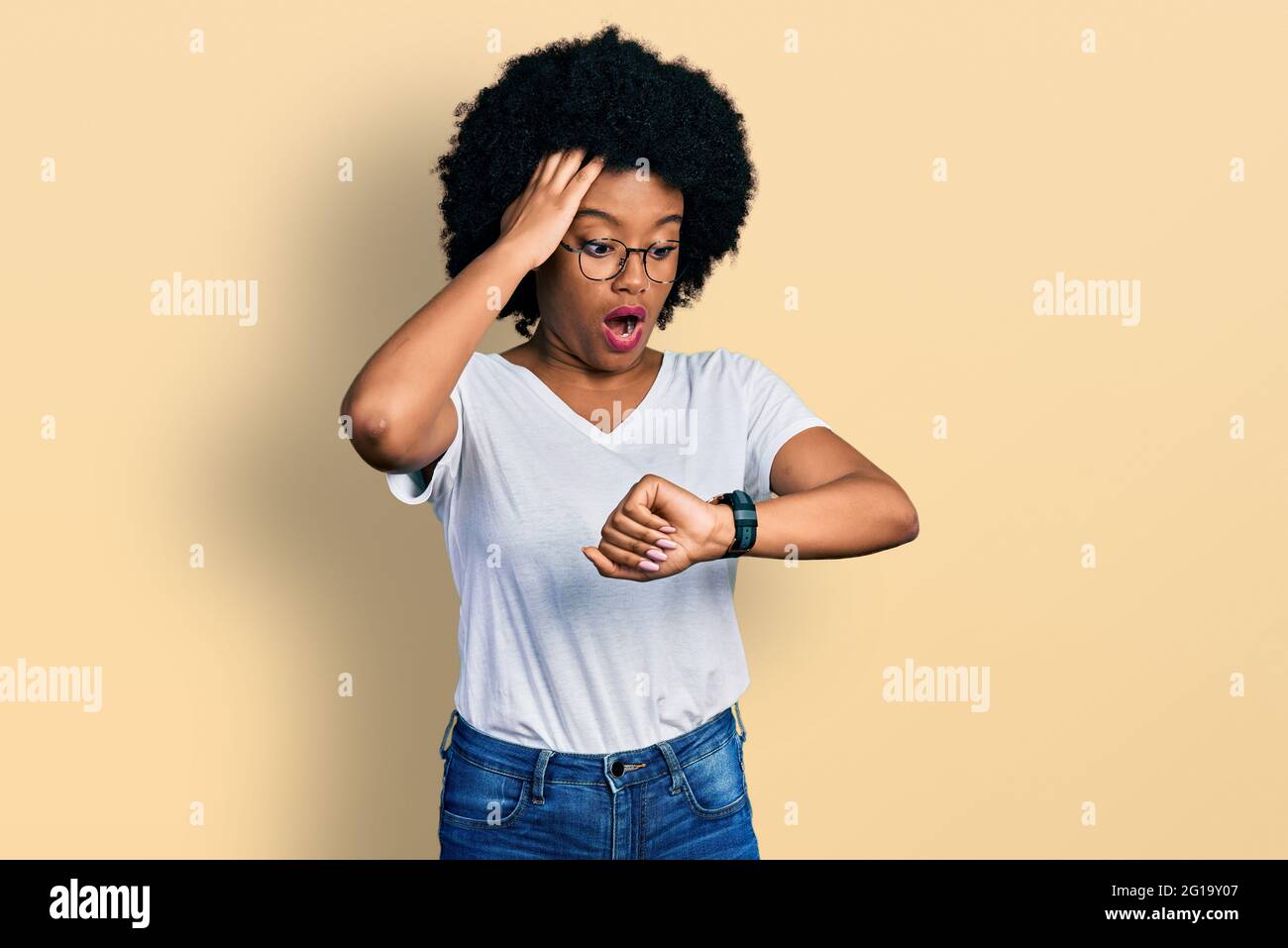 Junge afroamerikanische Frau, die ein legeres weißes T-Shirt trägt und besorgt auf die Uhr schaut, aus Angst, zu spät zu kommen Stockfoto