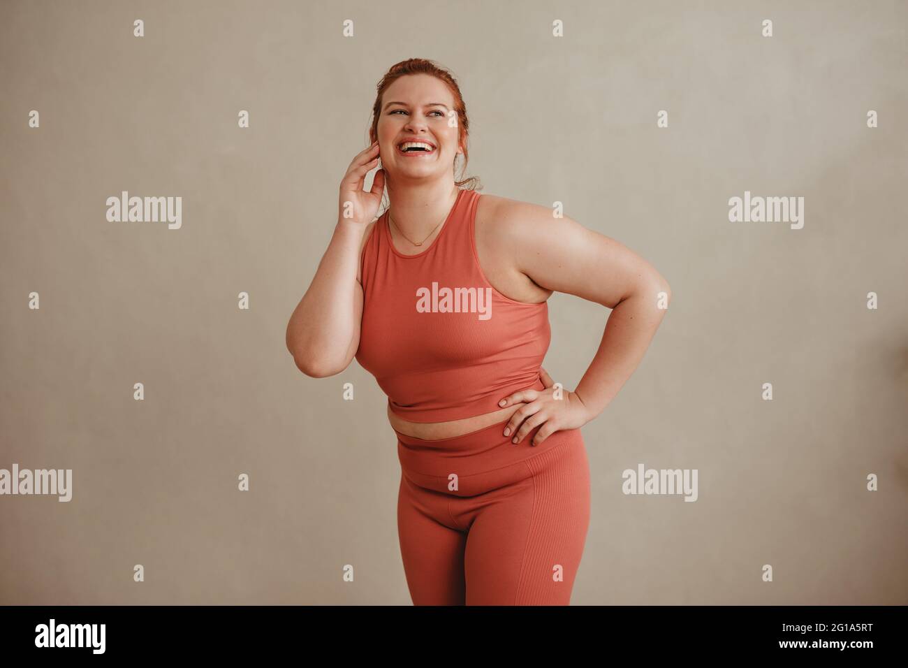 Selbstbewusste Frau in Sportkleidung, die wegschaut und lächelt. Gesunde und große Frau, die im Fitnessstudio gegen eine Wand steht. Stockfoto
