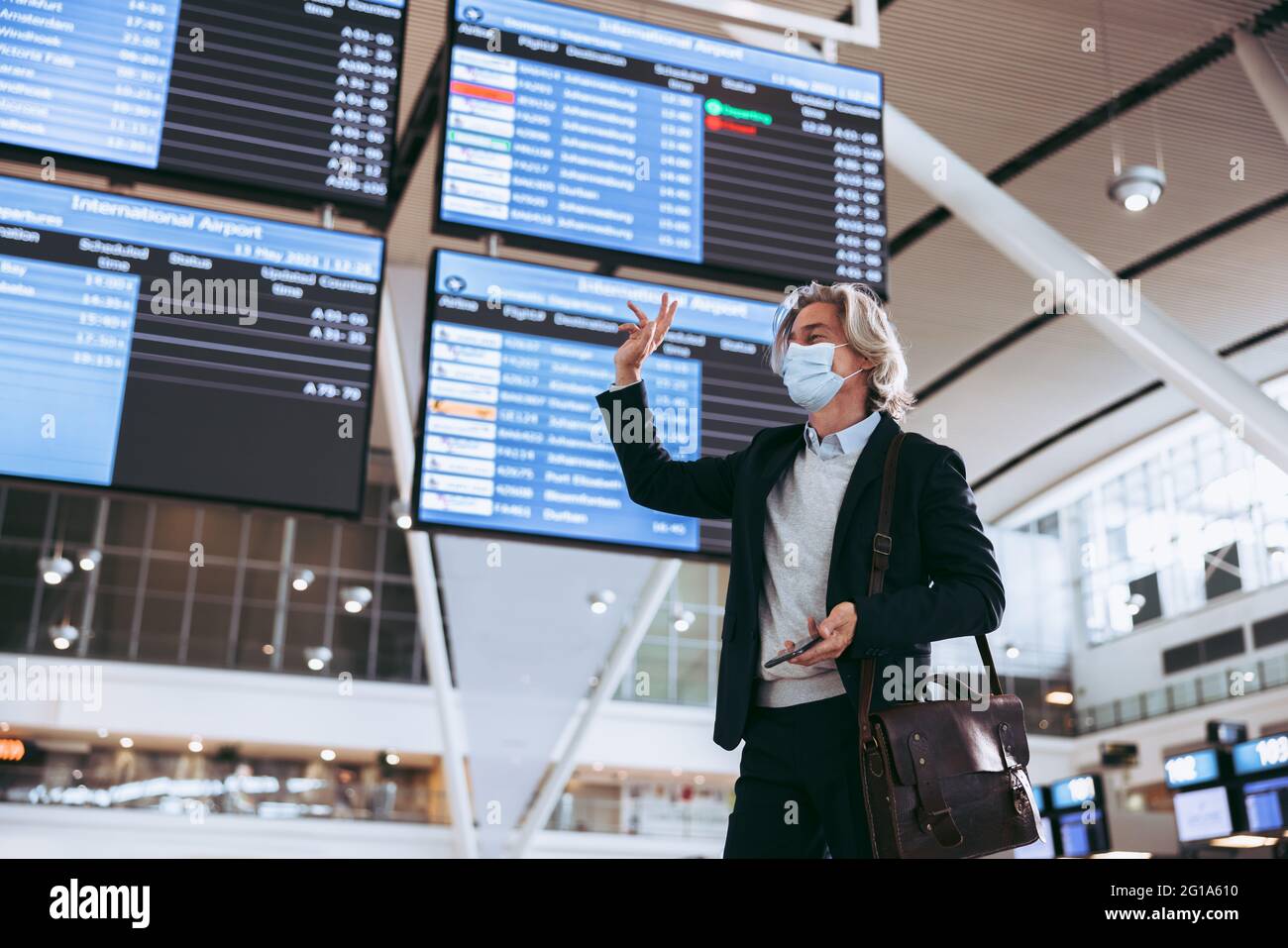 Mann mit Gesichtsmaske, der am Flughafenterminal steht und jemanden anwinkt. Geschäftsmann, der nach der Sperrung einer Pandemie reist. Stockfoto