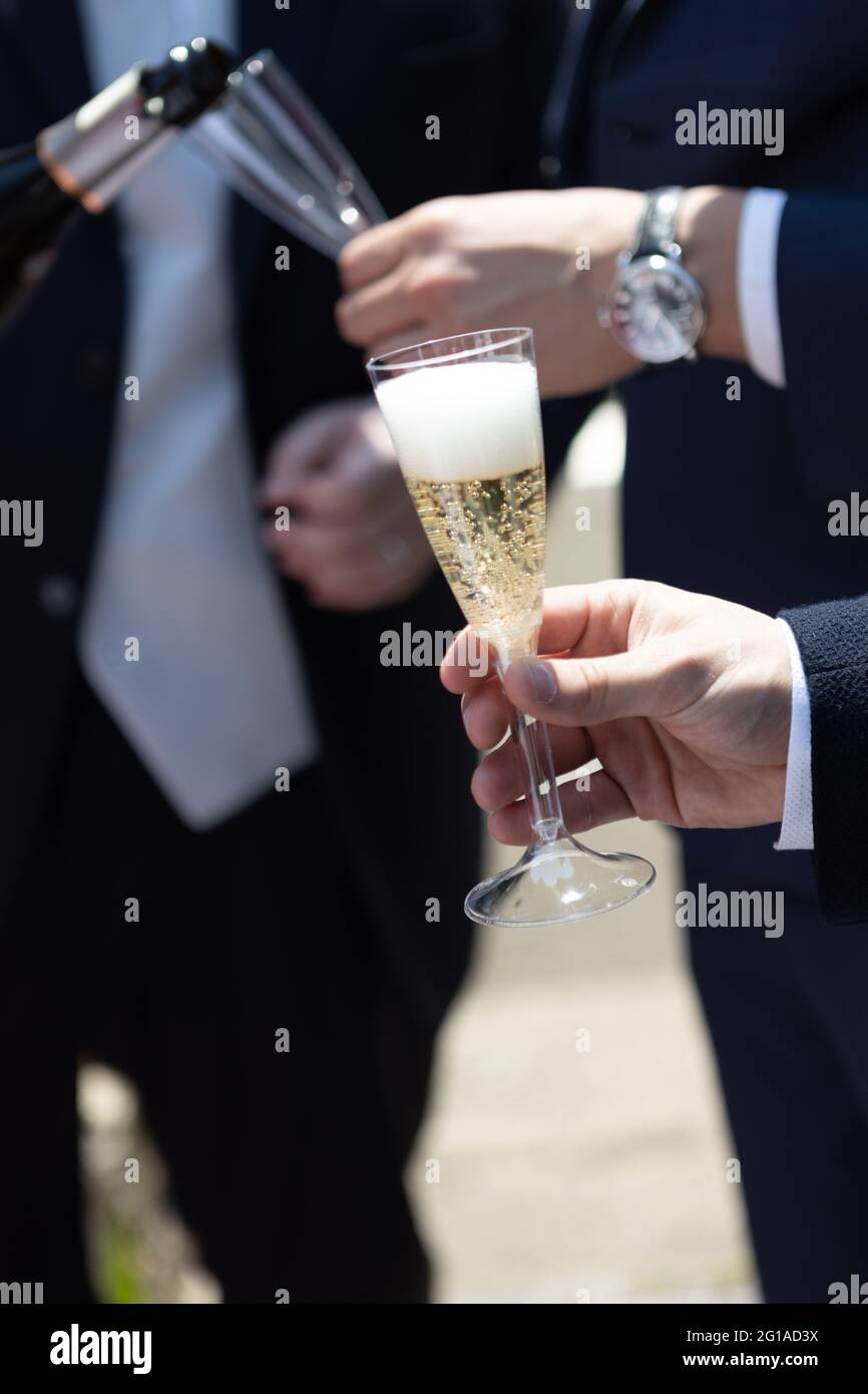 Zwei elegante Männer im Anzug, die Sekt in ein Kunststoffflötenweinglas gießen Stockfoto