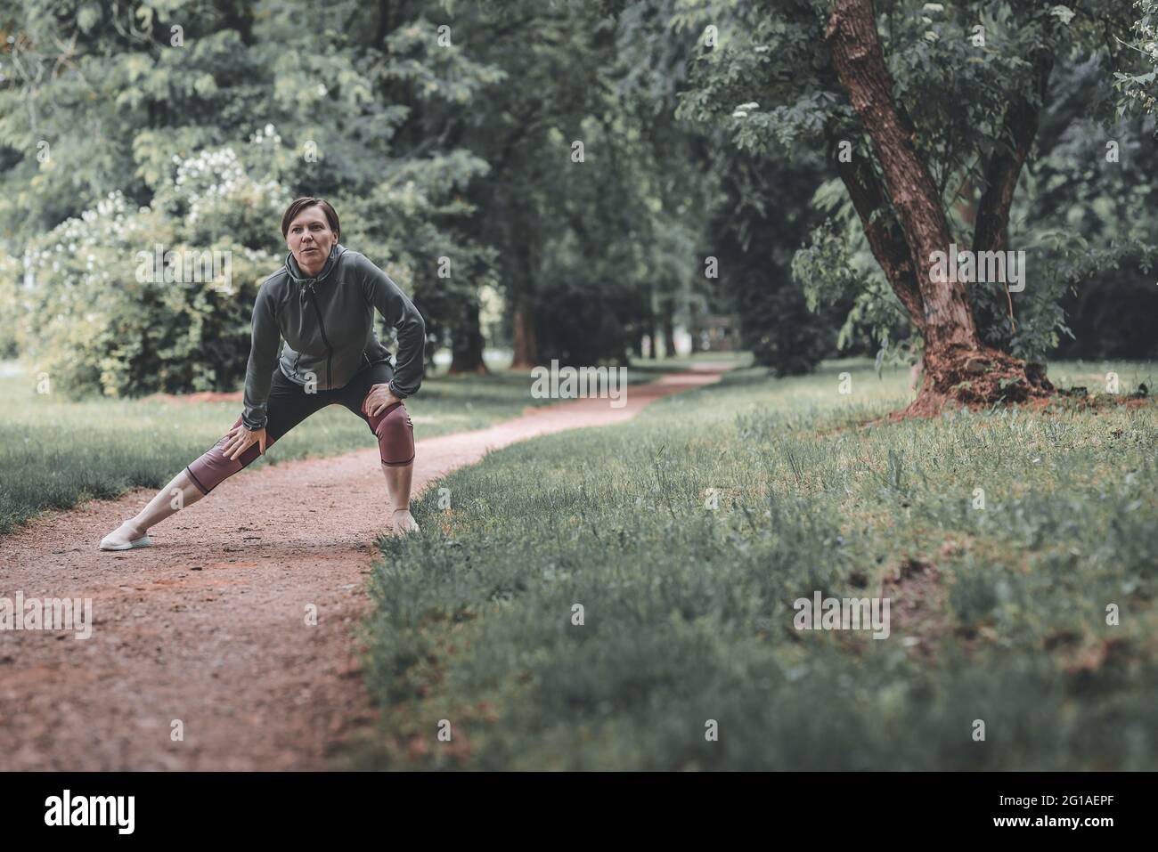 Porträt einer erwachsenen Joggerin, die sich in den 40er Jahren im Park dehnt und sich aufwärmt, um auf der Strecke zu laufen, gesund zu leben und sich zu sportlich zu betätigen Stockfoto