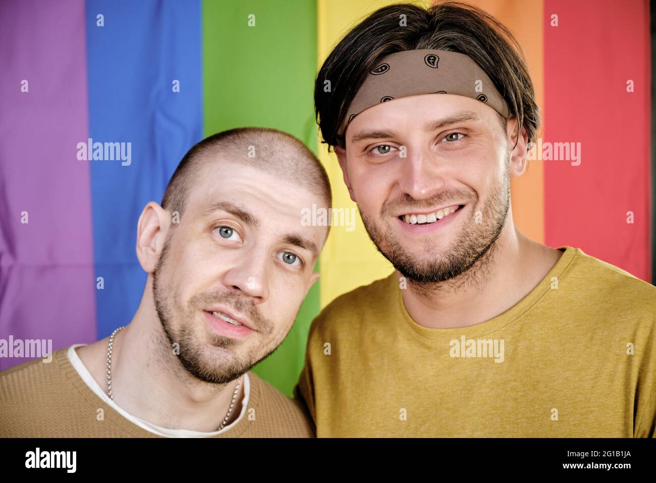 Zwei glückliche schwule Männer stehen gegen Regenbogenfahne und schauen dich an Stockfoto