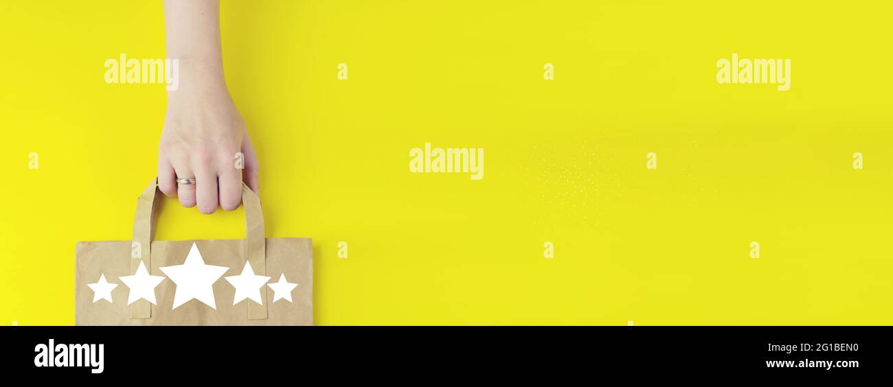 Customer Experience Konzept. Einkaufstasche aus recyceltem braunem Papier in der Hand mit Hologramm fünf Sterne 5 Rating-Symbol auf gelbem Hintergrund, flach liegend. Sommer s Stockfoto