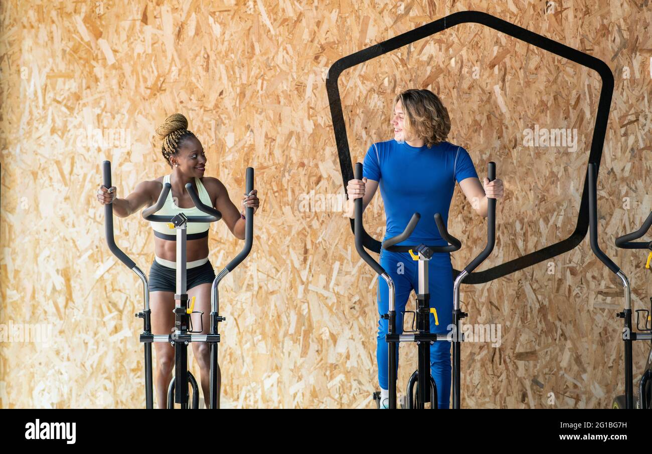 Lächelnde, multirassische Athleten in Sportkleidung trainieren auf Übungsfahrrädern, während sie sich in der Turnhalle gegenseitig anblicken und sprechen Stockfoto