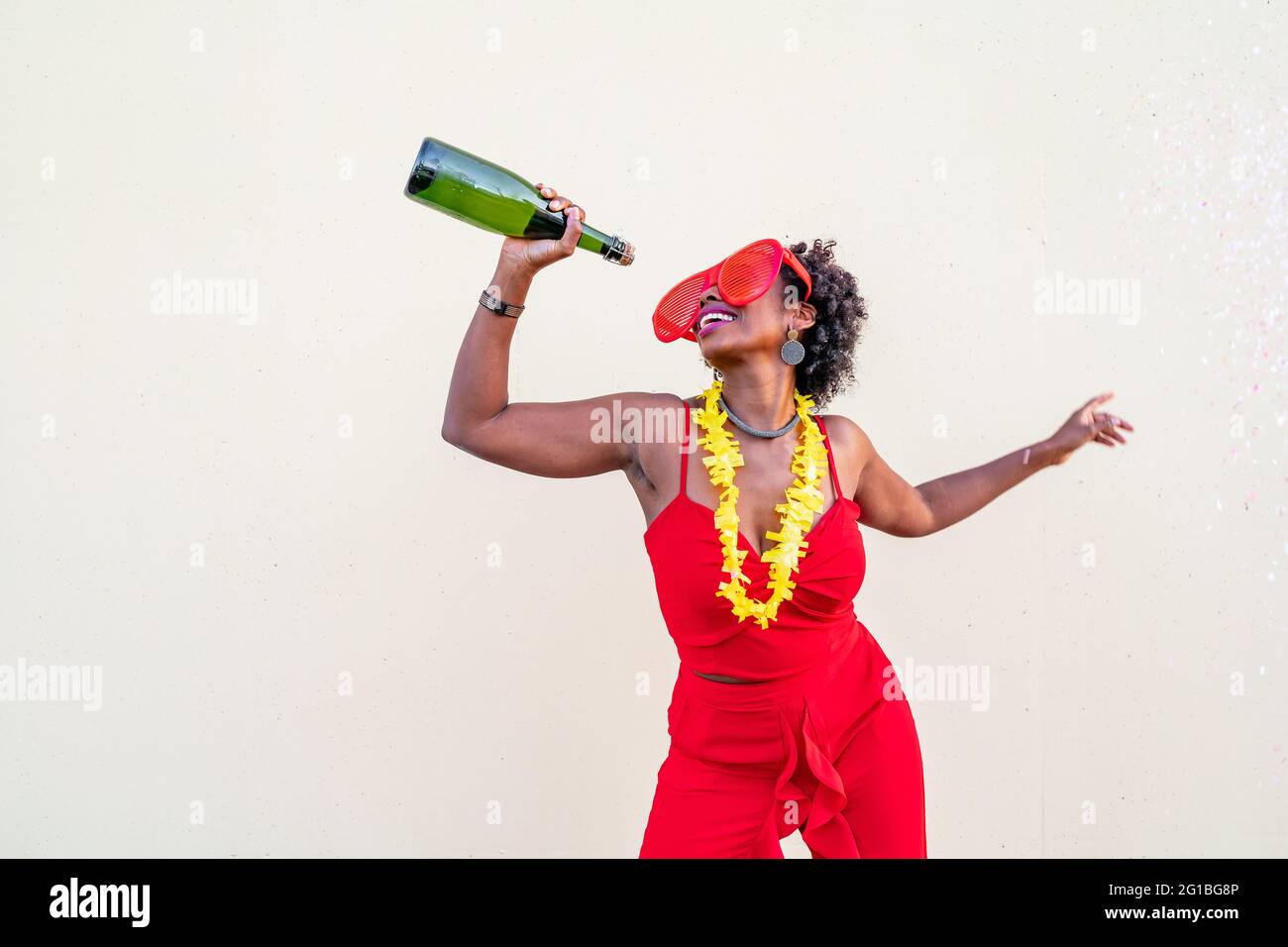 Fröhliche afroamerikanische Frau in roter Kleidung und Brille mit einer Flasche alkoholischen Getränks während der Party auf hellem Hintergrund Stockfoto