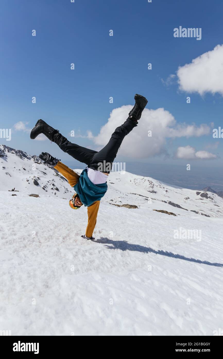 Anonymer Sportler in einer Brille, die mit erhobenen Beinen auf Schnee gegen die Sierra Nevada unter wolkig-blauem Himmel auf der Hand steht Stockfoto