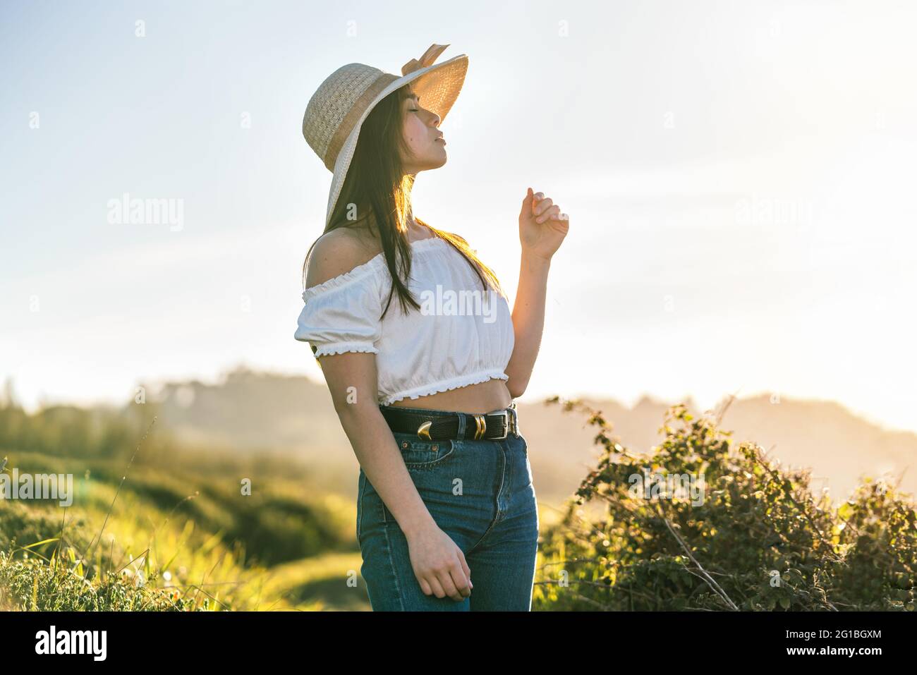 Attraktive, verträumte Hündin in Jeans und weißem Top und in angenehmen Gedanken wegschauen, während sie an sonnigen Tagen auf üppigem, grasbewachsenen Gelände steht Stockfoto