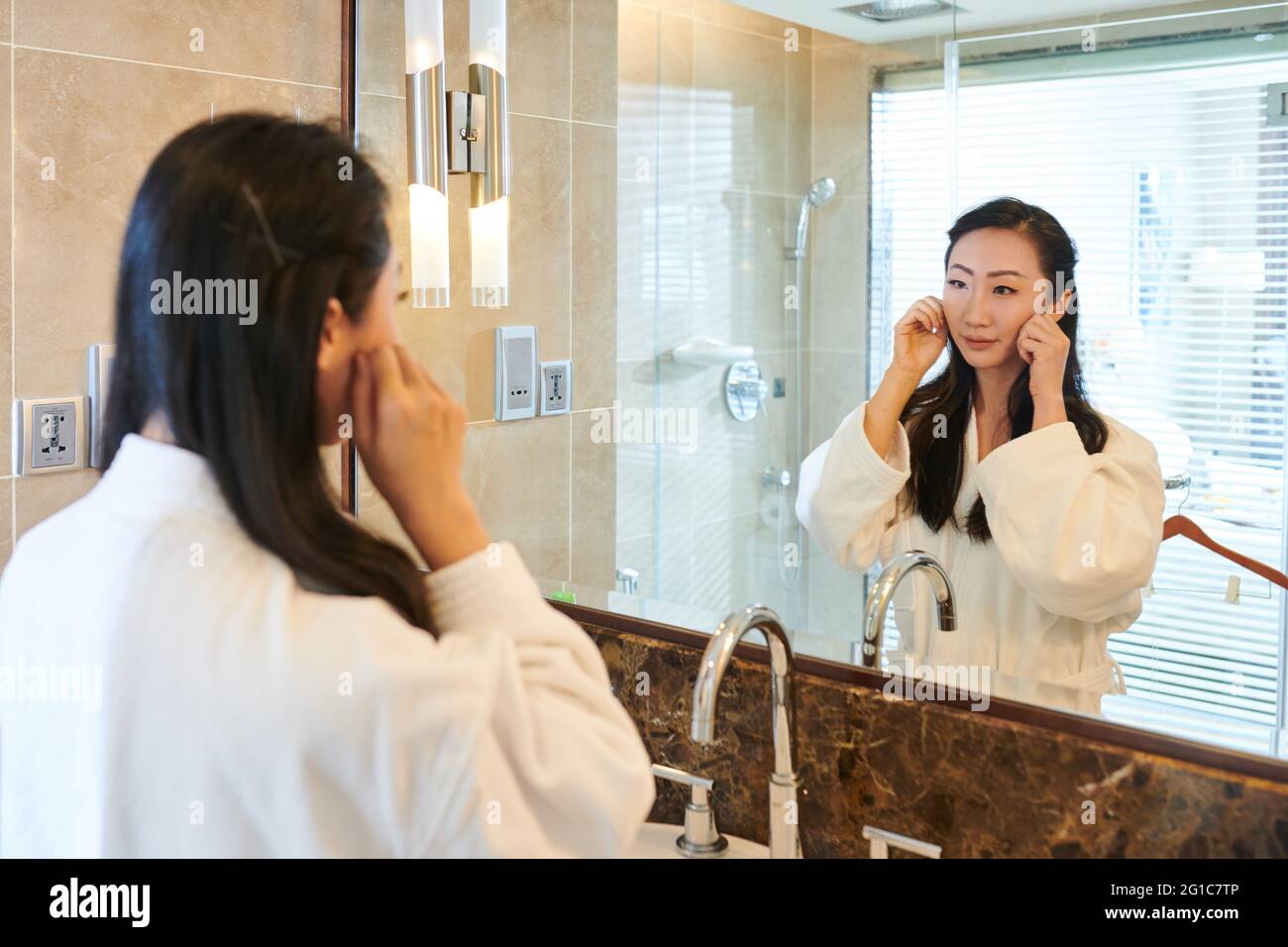 Hübsche junge Frau im Bademantel, die im Badezimmer vor dem Spiegel steht und ihr Gesicht massiert Stockfoto
