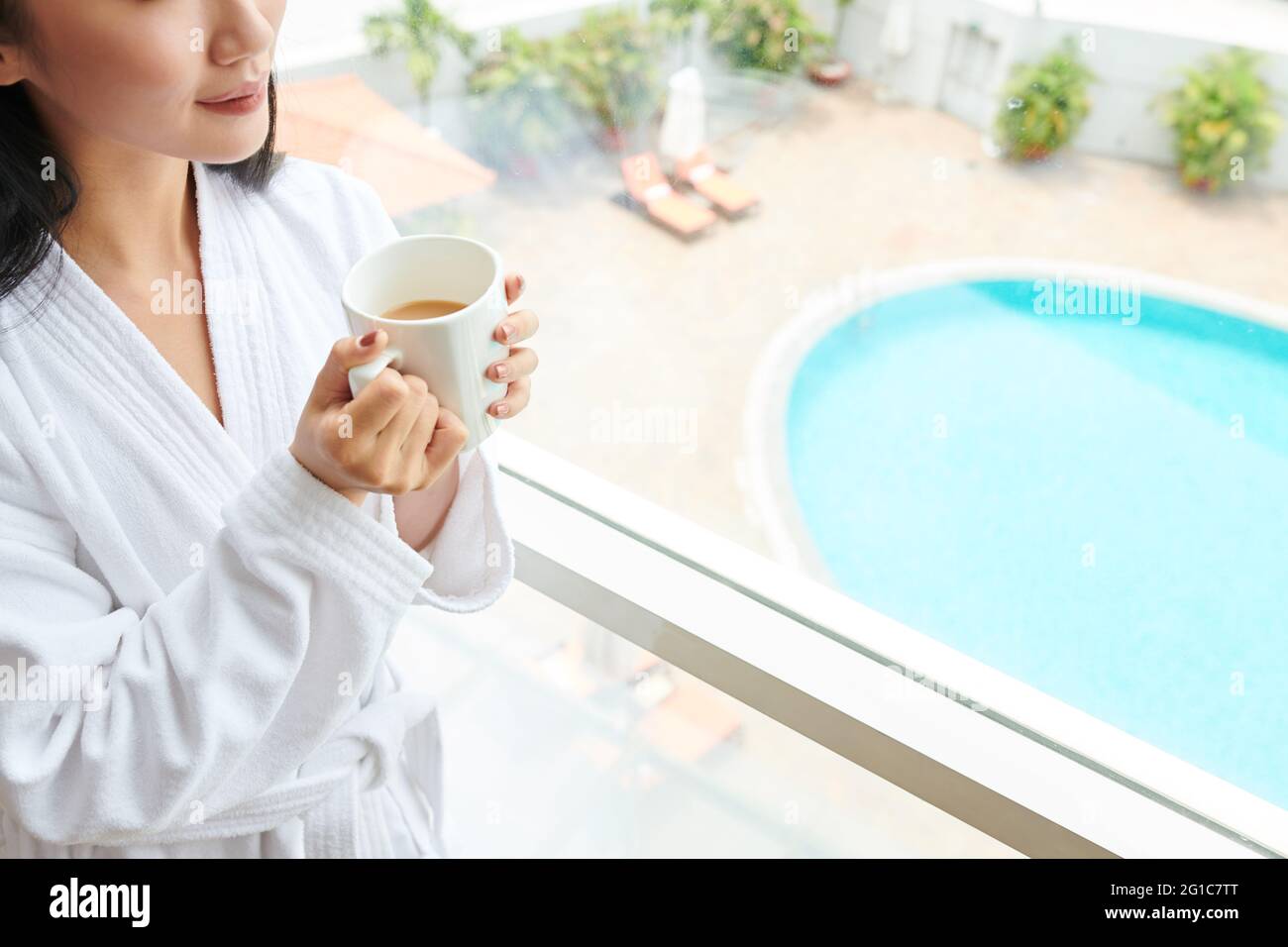 Beschnittenes Bild junge Frau im weißen Bademantel trinkt einen großen Becher Morgenkaffee, wenn sie am Hotelfenster steht und nach draußen schaut Stockfoto