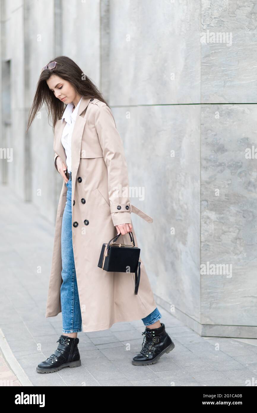 Ein junges hübsches Mädchen in Regenmantel und Jeans geht mit einer Handtasche in der Hand in der Nähe eines hohen Gebäudes durch die Stadt. Stockfoto
