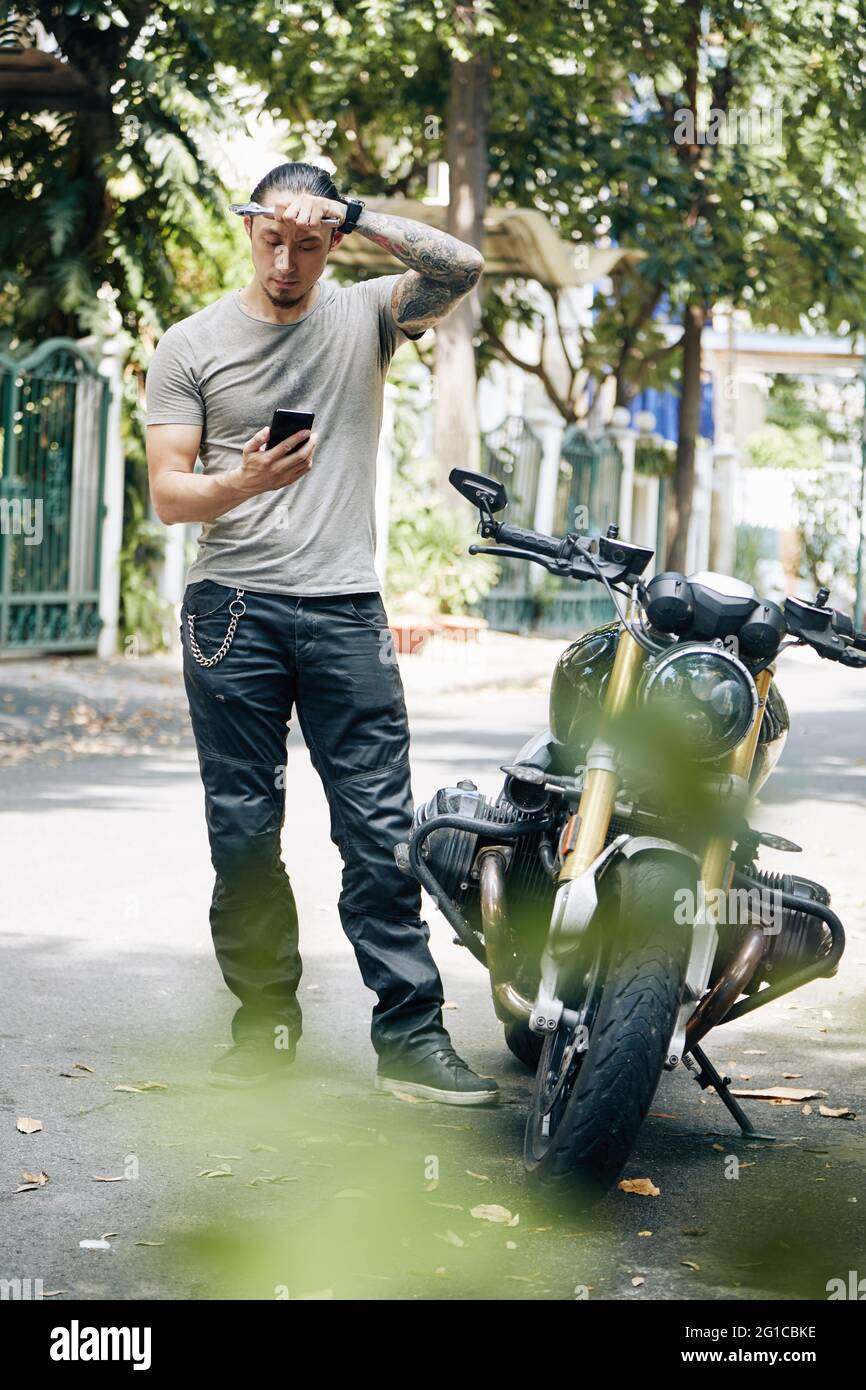 Verwirrt Mann mit Schraubenschlüssel in den Händen abwischen Schweiß von ihrer Stirn und lesen Artikel über Motorrad Reparatur auf seinem Smartphone Stockfoto