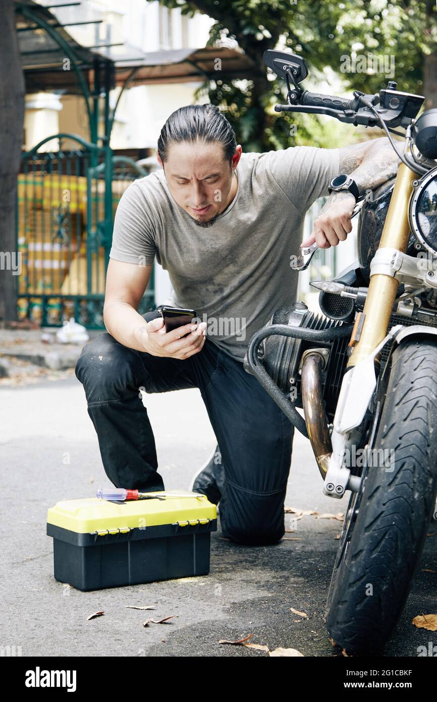 Frowning seriöse Mechaniker lesen Handbuch auf Smartphone-Bildschirm bei der Reparatur Motor seines Motorrads Stockfoto