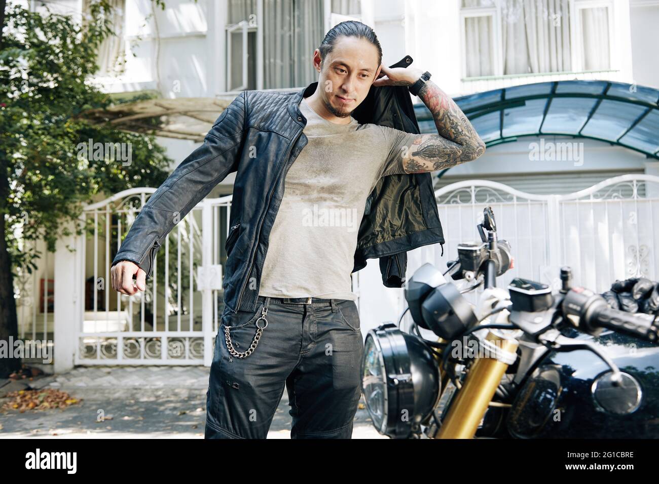 Schöner stilvoller Mann, der eine schwarze Lederjacke aufsetzt, wenn er bereit ist, auf dem Motorrad zu fahren Stockfoto