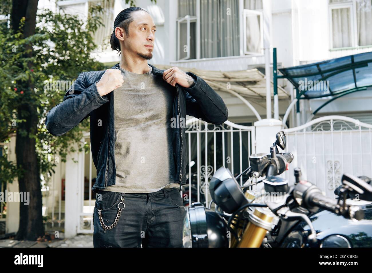 Nachdenklicher ernsthafter Motorradfahrer im verschwitzten T-Shirt, der beim Fahrrad steht und eine schwarze Lederjacke aufsetzt Stockfoto