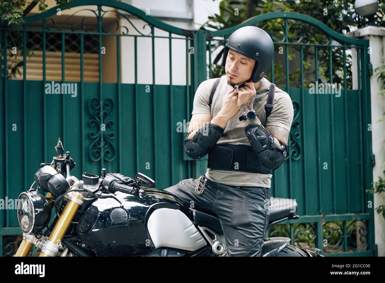 Pensiver Mann sitzt auf einem Motorrad, trägt Fahrausrüstung und T-Shirt und befestigt seinen Helm Stockfoto