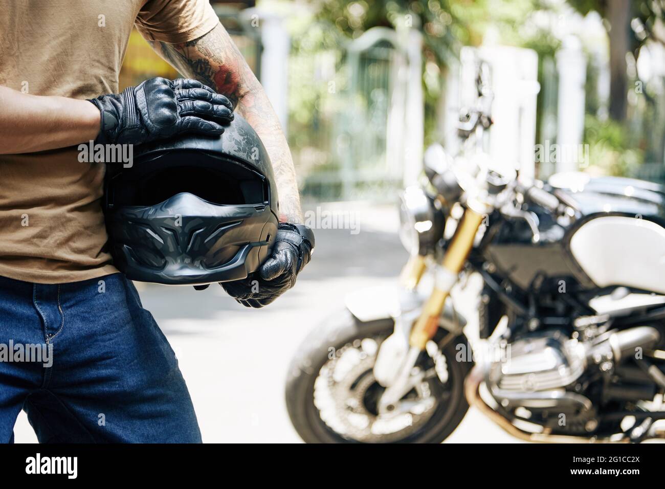 Zugeschnittenes Bild eines Motorradfahrers in Lederhandschuhen, der seinen Helm hält, sein Fahrrad ist im Hintergrund Stockfoto
