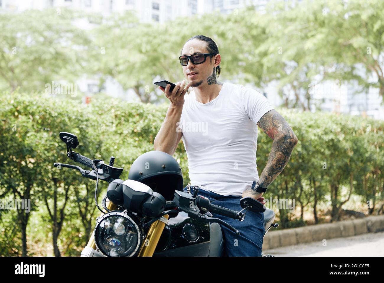 Mann in einer Sonnenbrille, der neben seinem Motorrad steht und eine Sprachnachricht für einen Freund oder Kollegen aufzeichnet Stockfoto