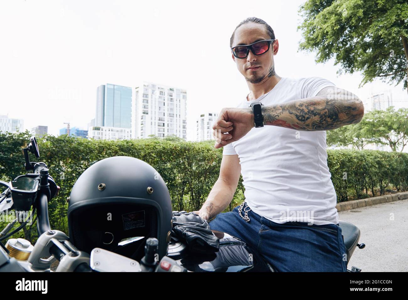 Mann mit Sonnenbrille und weißem T-Shirt, der auf dem Motorrad oder Roller sitzt und die Zeit oder Benachrichtigungen auf seiner Smartwatch überprüft Stockfoto