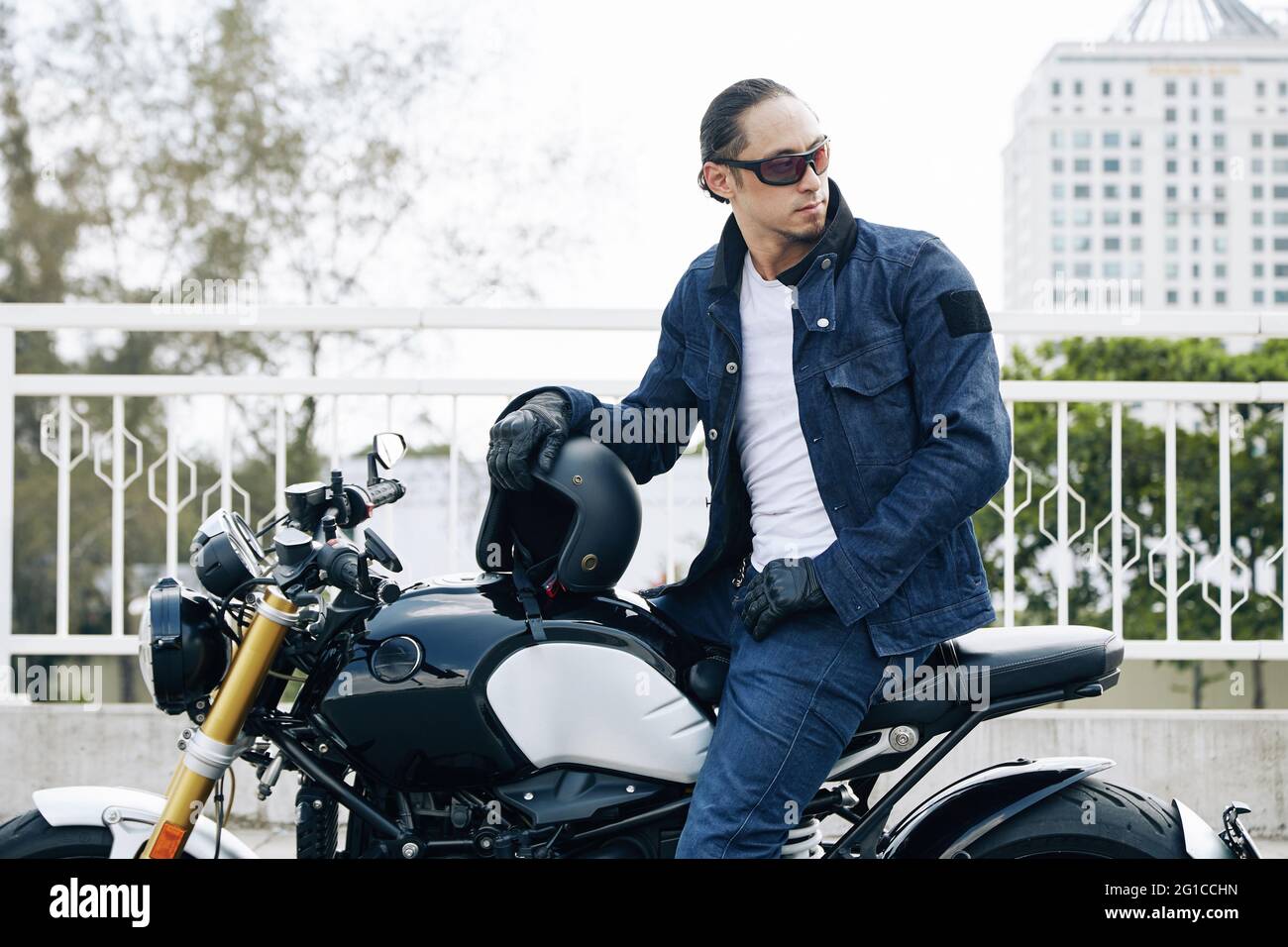 Stilvoller Mann in Sonnenbrille, Jeans und Jeansjacke sitzt auf dem Motorrad, zieht den Helm aus und schaut zurück Stockfoto