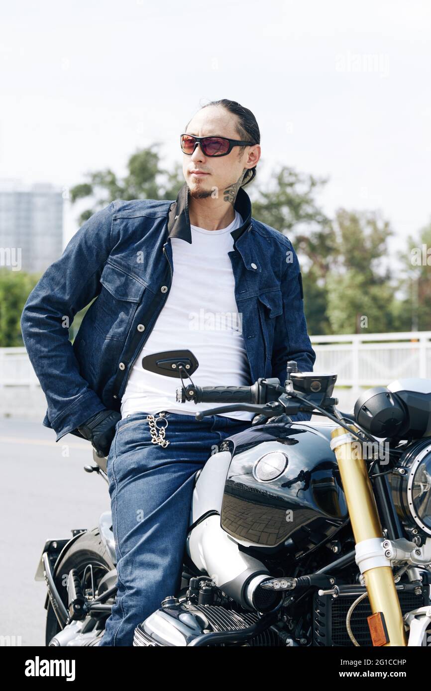 Porträt eines lächelnden, gutaussehenden Mannes in Denim-Jacke, der auf dem Motorrad sitzt und wegschaut Stockfoto