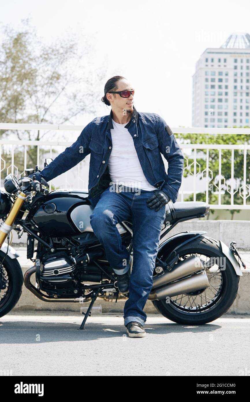 Hübscher, stylischer Motorradfahrer in Jeans und Jeansjacke, der sich am Motorrad lehnt und wegschaut Stockfoto
