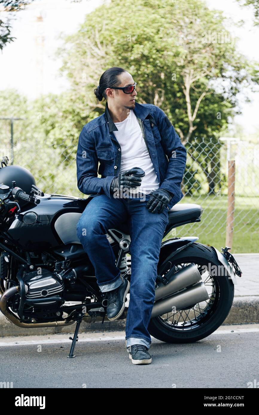 Schöner Mann in Jeans und Jeansjacke, der sich auf dem Motorrad lehnt und nach einer langen Fahrt eine Tasse Kaffee zum Mitnehmen trinkt Stockfoto