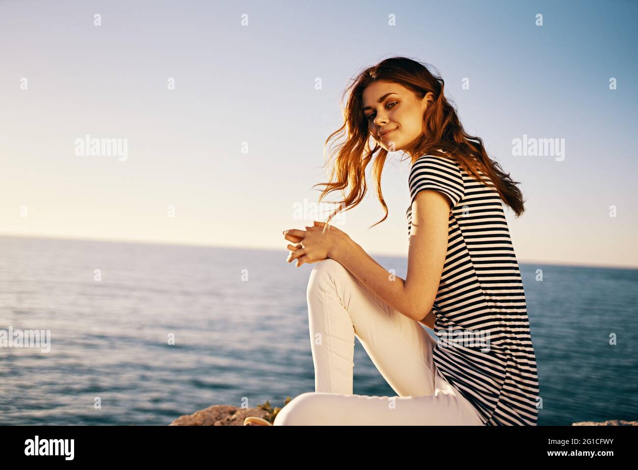 Eine Frau sitzt in einem gestreiften T-Shirt und einer weißen Hose Auf einem Stein in der Nähe des Meeres im Sommer Stockfoto