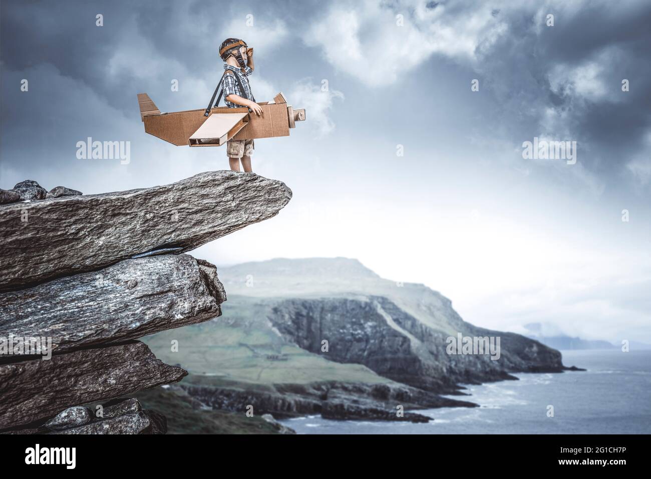 Kind, das als Flieger mit einem Flugzeug aus Pappe auf einer Klippe gekleidet ist, träumt von einem Abenteuer. Stockfoto