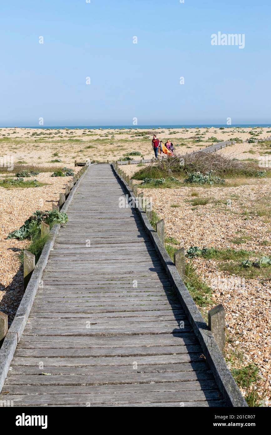 Ein Holzsteg über die Single zum Strand mit einer Familie, die zur Kamera in Dungeness, Kent, England, Großbritannien, geht. Stockfoto