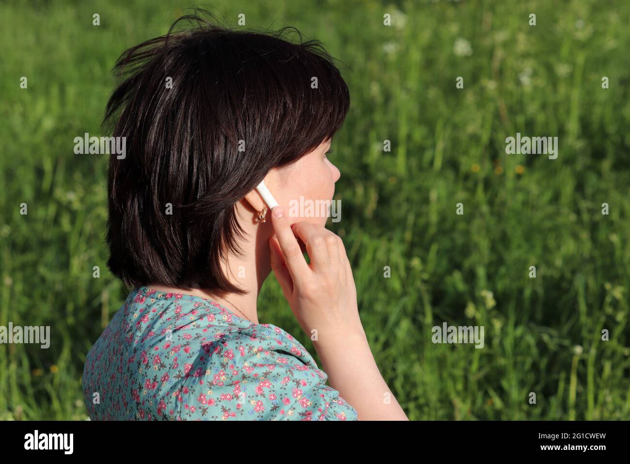 Die Frau berührt den kabellosen Kopfhörer im Ohr, das auf der Sommerwiese steht. Headset, Musik hören und Sprachanruf in einer Natur Stockfoto