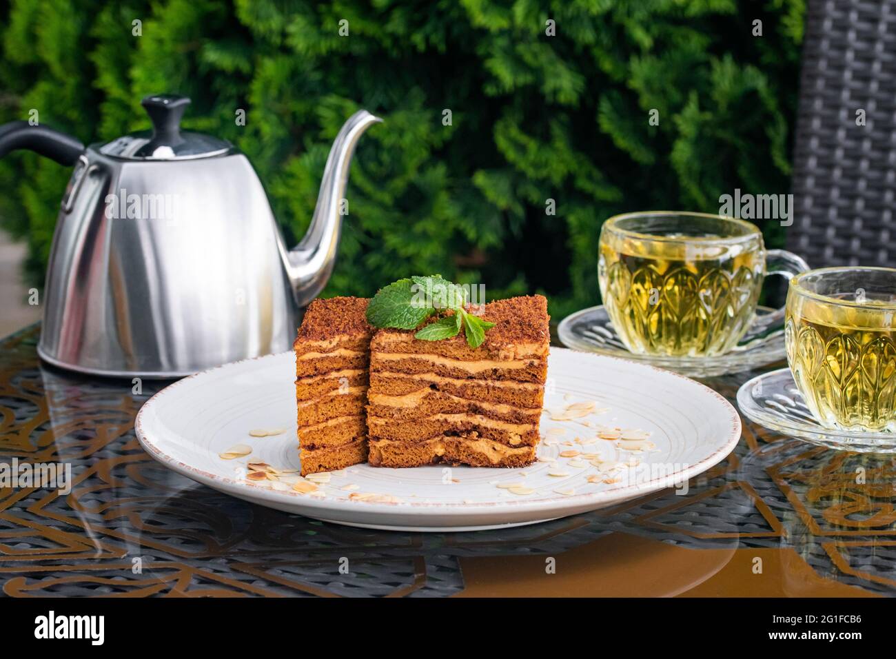 Medovik Kuchen mit Krümel bedeckt, zwei Tassen mit Minztee und Stahlkessel auf dem Tisch im Sommercafe. Teetrinkkonzept, britische Kultur. Stillleben. Stockfoto