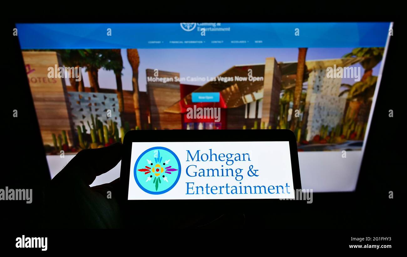 Person, die das Smartphone mit dem Logo des US-amerikanischen Unternehmens Mohegan Gaming and Entertainment (MGE) auf dem Bildschirm vor der Website hält. Konzentrieren Sie sich auf die Telefonanzeige. Stockfoto